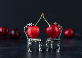 Couple of red ripe cherries on two vintage small silver chairs with blurred cherries in the black backround with copy space