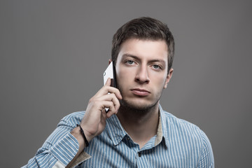 Moody portrait of confident young stubble man in blue shirt talking on the cellphone looking at camera