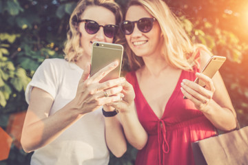 Sunny summer day. Close-up of a smartphone in the hands of young women standing outdoors. Girl in white t-shirt shows a picture on the screen of the smartphone girl in a red dress. Girls using gadget.