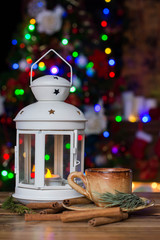 Christmas Decorations with small cup of coffee, lamp with candle, cinnamon sticks, fir branch on wooden table against lights background