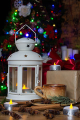 Christmas Decorations with small cup of coffee, gift box, lamp with candle, cinnamon sticks, star anise, fir branch on wooden table against lights background