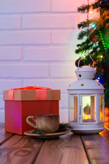 Christmas Decorations with cup of coffee, orange gift box, lamp with candle, fir branch on wooden table against brick background