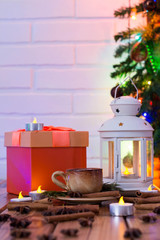 Christmas Decorations with cup of coffee, orange gift box, lamp with candle, cinnamon sticks, star anise, fir branch on wooden table against brick background