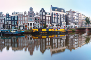 Fototapeta premium Panorama kanału Amsterdam Singel z typowymi holenderskimi domami, mostem i łodziami mieszkalnymi w porannej niebieskiej godzinie, Holandia, Holandia.