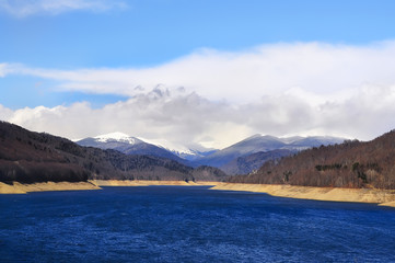 blue lake at the foot of the mountains covered with snow, beautiful clouds and sky, the forest slopes. Beautiful winter landscape, the beginning of spring.