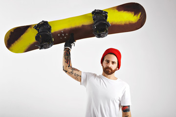 Serious athletic young man with tattoos and beard raising his snowboard with one hand isolated on white