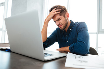 Confused young man sitting near laptop and holding head
