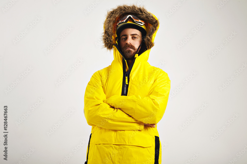 Wall mural Attractive young man wearing snowboard helmet and goggles and a bright yellow anorak with arms crossed on white background