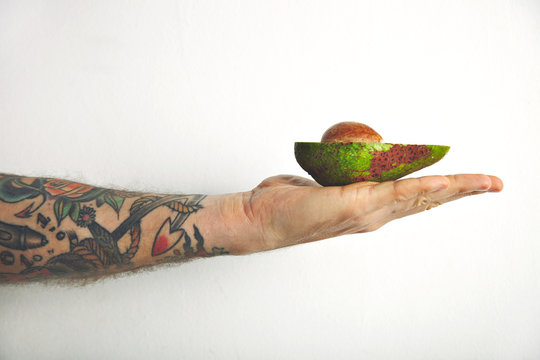 Half of an avocado with stone inside lying on a hand of a man with tattoos, close up shot isolated on white