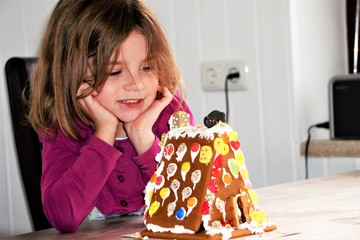 Fototapeta premium Ein kleines Mädchen sitzt am Küchentisch in der Küche vor einem selbstgemachten Lebkuchenhaus und freut sich.