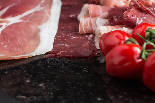 Ham mix. jambon. Traditional Italian and Spanish salting, smoking, dry-cured dish - jamon Serrano and prosciutto crudo sliced with herbs and tomatos on dark stone background. Copy space. Closeup. 