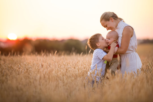 Happy family enjoying sunset in wheat field. Beautiful young woman with adorable baby boy and kid boy. Mother hugging her two children on a meadow on a sunny evening. Mom and sons. Outdoors.