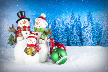 Christmas decoration with snowman figurines as a family, colorful baubles on a blue snow pine tree landscape background.