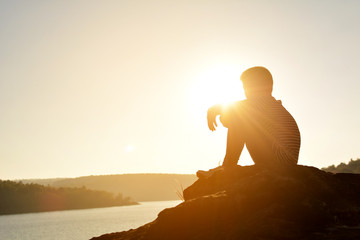 Silhouette of sad boy and sitting on the rock in river at the sky sunset