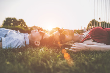 Summer sunny evening. Backlight. Two young woman in sunglasses lying on grass heads together. One...