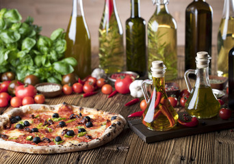 Delicious, tasty and fresh, rustic Italian pizza, served on wooden table. Bunch of tomatoes, chili, basil, garlic in background. Bowls with different kinds of pepper. Many bottles of olive oil