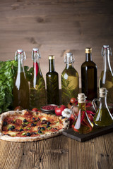 Delicious, tasty and fresh, rustic Italian pizza, served on wooden table. Bunch of tomatoes, chili, basil, garlic in background. Bowls with different kinds of pepper. Many bottles of olive oil