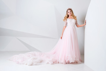 Beautiful blonde woman wearing gorgeous long dress and crystal crown posing in studio on white polygonal background.