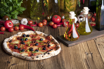 Delicious, tasty and fresh, rustic Italian pizza, served on wooden table. Bunch of tomatoes, chili, basil, garlic in background. Bowls with different kinds of pepper. Many bottles of olive oil