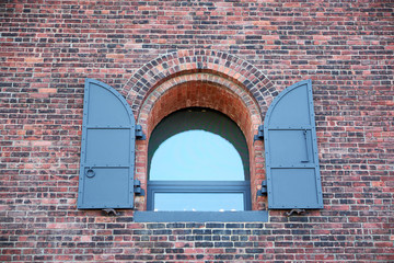 Arched window with belonging shutters on massive hinges, fastened to a brick facade of a former...