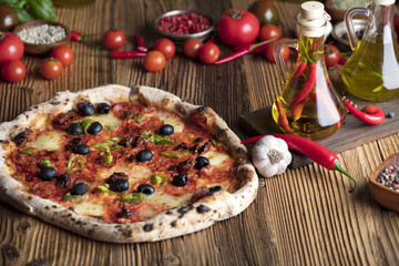 Delicious, tasty and fresh, rustic Italian pizza, served on wooden table. Bunch of tomatoes, chili, basil, garlic in background. Bowls with different kinds of pepper. Many bottles of olive oil
