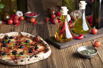 Delicious, tasty and fresh, rustic Italian pizza, served on wooden table. Bunch of tomatoes, chili, basil, garlic in background. Bowls with different kinds of pepper. Many bottles of olive oil