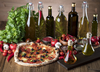 Delicious, tasty and fresh, rustic Italian pizza, served on wooden table. Bunch of tomatoes, chili, basil, garlic in background. Bowls with different kinds of pepper. Many bottles of olive oil