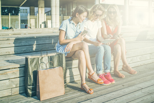 Summer sunny day, three young women resting after shopping sitting outdoors.In hands of girls smartphones and tablet computer.Nearby are brown and black shopping bags.Girls showing pictures on gadget.