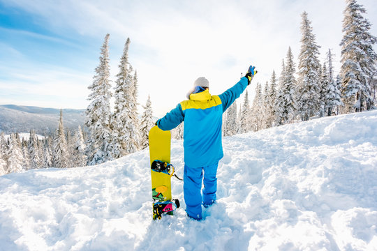 Snowboarder stands with snowboard on the top of mountain on forest backdrop on sunny winter day