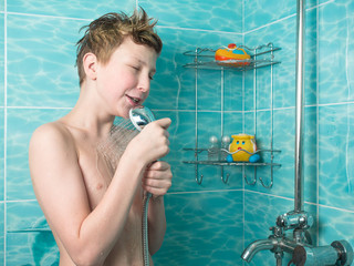 Young boy with red hair and naked torso holding a watering can shower and sings on the background of blue tiles and bottles of shampoo