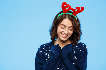 Young beautiful brunette girl in knited sweater and christmas reindeer antlers smiling  over blue background.