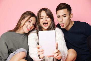 Young beautiful friends making selfie on tablet over pink background.