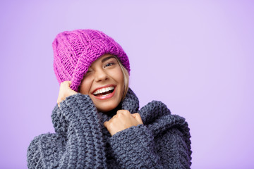 Young beautiful fair-haired girl in knited hat and sweater smiling winking looking at camera over violet background. 