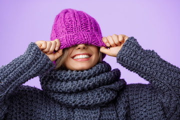 Young beautiful fair-haired girl in knited hat and sweater smiling over violet background. 