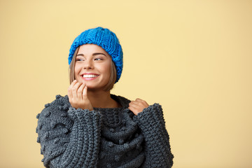 Young beautiful fair-haired girl in knited hat and sweater smiling looking at side over yellow background.