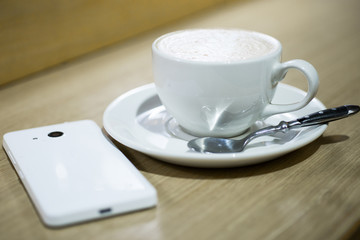 Coffee cappuccino in white cup and white mobile phone on the table in a cafe.
