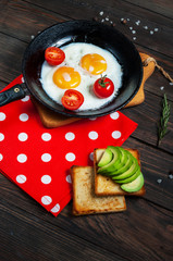 Pan of fried eggs and cherry-tomatoes with bread, avocado on dark wood table.