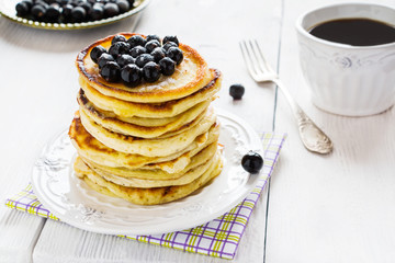 Stack of homemade pancakes with black currant, vintage white plate and fork, cup of coffee, wooden table.