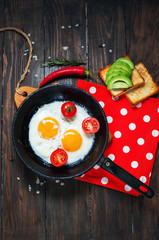 Pan of fried eggs and cherry-tomatoes with bread, avocado on dark wood table, top view