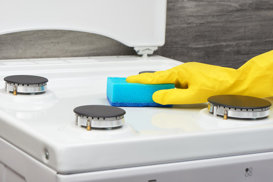 Hand in yellow glove cleaning white stove with blue sponge