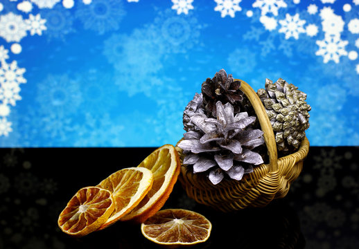 Christmas and New Year`s natural organic decorations with silver pine cones, dry orange fruits on a black reflection surface and blue snowy sky bokeh background with snowflakes.