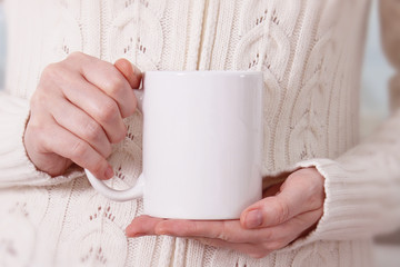 Girl in warm sweater is holding white mug in hands.