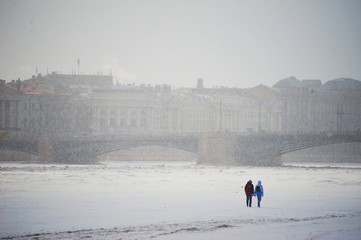 Two people walk on ice of Neva river in winter, in the background through the fog the bridge is visible and the architectural ensemble of the St. Petersburg waterfront.