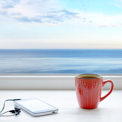Cup of coffee, smartphone and headphones on a windowsill. In the background sea and sky