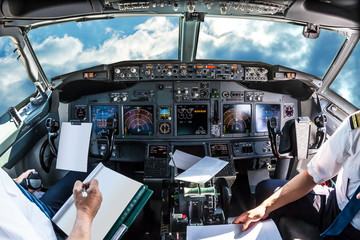 Airplane cockpit flying in a cloudy blue sky at day. with pilots arms and blank white papers for copy space. - obrazy, fototapety, plakaty