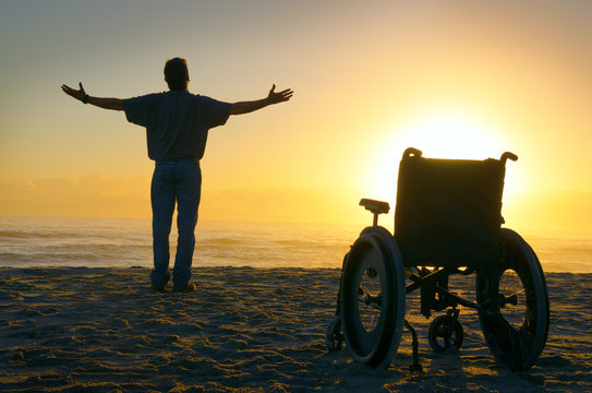 Miracle spiritual healing crippled man exalted arms spread at the ocean shoreline as he stands up out of his wheelchair and walks towards the sunrise