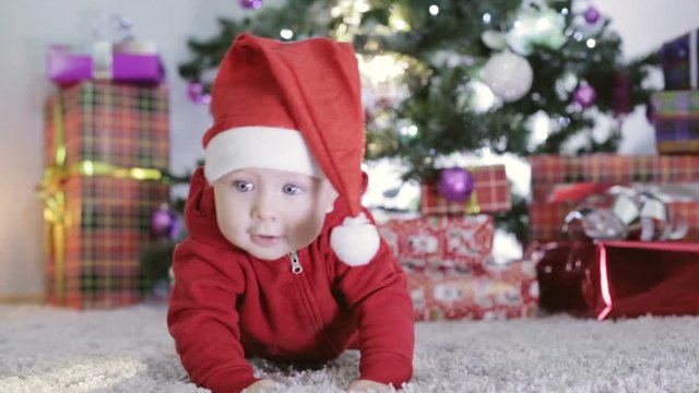 Baby boy in Santa costume crawling near Christmas tree.
