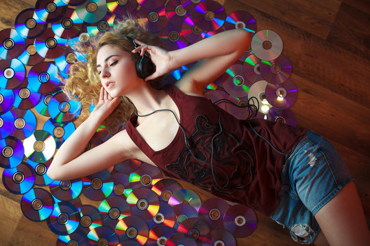 Concept: hipster lifestyle, relaxation. Beautiful young woman in headphones have fun and listen music lying on colorful CD's.