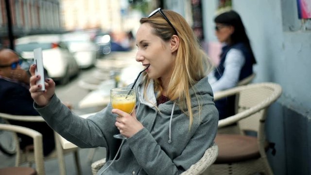 Happy girl doing selfies on smartphone while sitting outdoors and drinking juice
