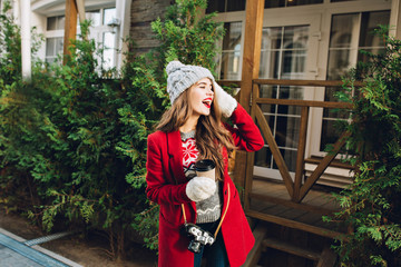Pretty young girl with long hair in red coat and knitted hat on wooden house  background. She holds coffee to go in white gloves, expressing to side